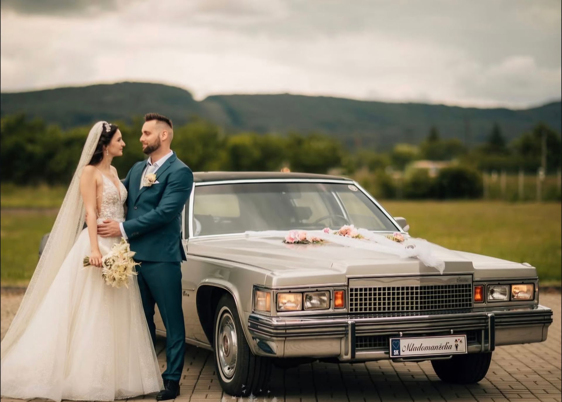 Novomanželia pri prednej časti Cadillacu Fleetwood v prírode s horami v pozadí